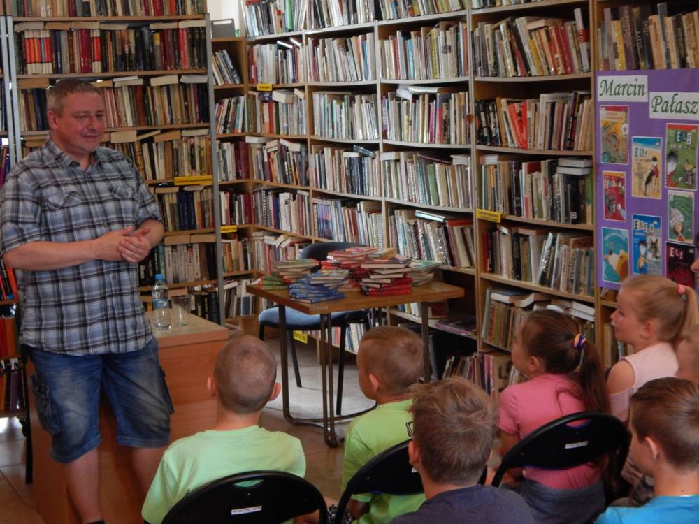 Biblioteka publiczna. Marcin Pałasz stoi przed uczniami. W tle regały z książkami.