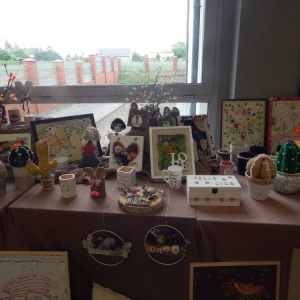 Prace mieszkańców DPS ułożone na stole, obrazki w ramkach, kaktusy z materiału, dekoracyjna szkatułka, kwiaty z włóczki